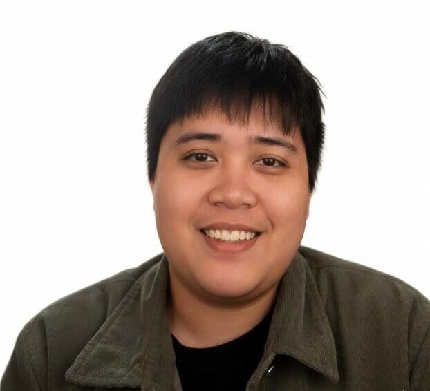 A person with short black hair is smiling at the camera, wearing a dark jacket over a black shirt, with a plain white background.